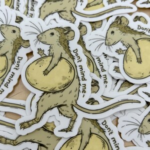 May include: A collection of stickers featuring a cartoon rat carrying a large, round cheese. The rat is light brown with a long tail and the text "Don't mind me-" is printed on the sticker. The cheese is yellow.