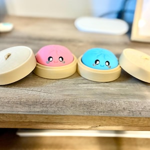 May include: Two colorful, smiling dumpling-shaped objects, one pink and one blue, sit in bamboo steamer baskets. The dumplings have cute, cartoon-like faces. Two matching lids are also present, creating a playful, food-themed design.