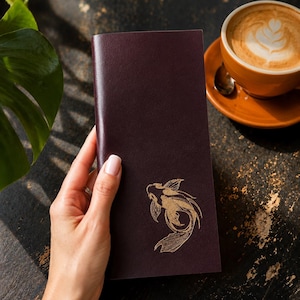Può includere: Una copertina per taccuino rettangolare in pelle color bordeaux scuro con un'illustrazione di un pesce koi impressa in oro sulla parte anteriore. Il taccuino presenta una texture liscia e opaca e un design pulito con angoli arrotondati.