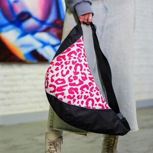 May include: A black and white sling bag featuring a pink leopard print. The bag has a long black strap and a curved shape. The bag is held by a person wearing a grey sweatshirt and a denim skirt.
