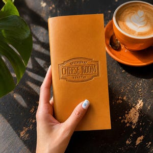 May include: A rectangular tan leather menu cover featuring a debossed emblem with the text RESTAURANT CHEESE ROOM SEATTLE centred on the front. The cover has a smooth, matte finish and is held vertically against a dark, textured surface.