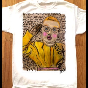 Op de afbeelding: Wit T-shirt met een gele en roze illustratie van een persoon met een bril en een geel pak. De tekst "THEY TELL US THAT WE LOST OUR TAILS-EVOLVING UP FROM LITTLE SNAILS-IS SAY THAT'S ALL JUST WIND IN SAIL-SARE WE-DEVO-WE'RE FINHERES-WE ARE NOT WHOLE PINHEADS ALL MONKEY IN * BUSWESSO SUITS TRACHERS MEN WEAR NOWNE WE'RE HOMO MEN GOD MAN MADE BUT HE MONKEY +0 DO ARE... DEVO WE MUST REPEAT... FARE WE NOT MEN? WE ARE ALL DEVO" is op het T-shirt gedrukt.