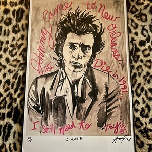 Peut inclure: Un dessin en noir et blanc d'un homme aux cheveux noirs et à l'expression sérieuse. Le dessin est sur un fond beige avec du texte rouge qui dit "Come to New Orleans Die in 1991 I still need to try". Le bas du dessin est signé "L.A.M.F. A/123".