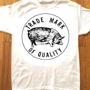 May include: White t-shirt with a black and white graphic of a pig inside a circle with the text "Trade Mark of Quality".
