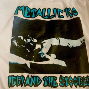 以下が含まれることがあります： 「Iggy and the Stooges」のバンドの青い黒のグラフィックプリントが施された白いTシャツ。画像の上に「METALLICKO」というテキストがあります。