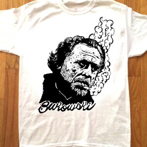 以下が含まれることがあります： 白地に黒と白のイラストがプリントされたTシャツ。イラストは、ひげを生やした男性と「Bukowski」という文字。