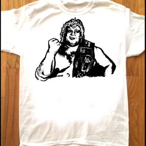 May include: A white t-shirt with a black and white illustration of a wrestler holding a championship belt. The wrestler is wearing a vest and has a mustache.