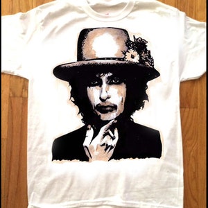 Puede incluir: Camiseta blanca con una ilustración en blanco y negro de una persona con un sombrero con flores. La persona está mirando hacia abajo y tiene una mano en la barbilla.
