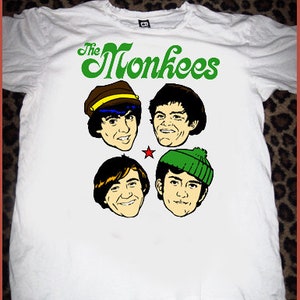 May include: White t-shirt featuring the text "The Monkees" in green, with cartoon illustrations of the band members' faces. The illustrations include a star and the band members wearing hats. The shirt is on a leopard print background.