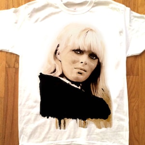 May include: White t-shirt featuring a sepia-toned portrait of a woman with blonde hair and a black top. The image is centered on the shirt, with the background being a wooden surface.