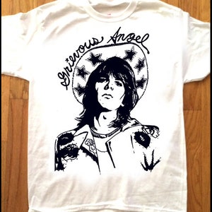 Könnte beinhalten: Weißes T-Shirt mit einer Schwarzweißillustration einer Person mit langen Haaren, die eine Jacke und einen Heiligenschein mit Sternen trägt. Der Text "Grievous Angel" steht über dem Kopf der Person.