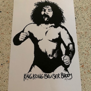 May include: Black and white illustration of a wrestler with a beard and long hair. The wrestler is shirtless and wearing wrestling trunks. The text "King Kong Bruiser Brody" is written below the wrestler.