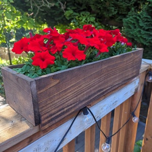 May include: A wooden planter box with red flowers hanging on a wooden railing with string lights.
