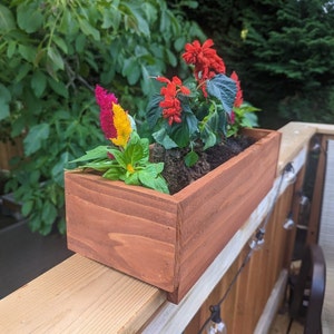 May include: A wooden planter box with a reddish-brown stain, filled with a variety of colorful flowers, including red, yellow, and pink. The planter is mounted on a wooden railing.
