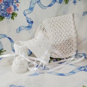 May include: White crocheted baby booties and bonnet set. The booties have a lace pattern and the bonnet has a scalloped edge.