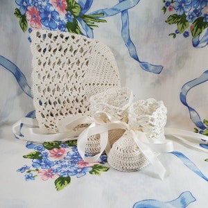 May include: A set of crocheted baby booties and a bonnet in white. The booties have a delicate lace trim and the bonnet has a scalloped edge.