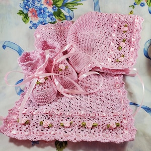 May include: A pink crocheted baby outfit with a matching hat and booties. The outfit is decorated with pink satin ribbon and small green fabric flowers.