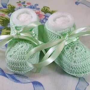 May include: A pair of light green crocheted baby booties with white satin ribbon laces. The booties are tied with a bow and are sitting on a white background with blue floral designs.