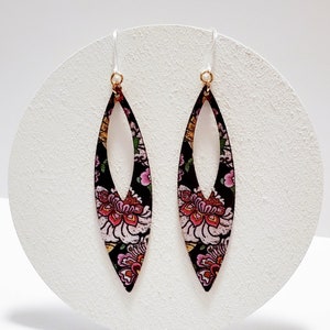 May include: A pair of long, teardrop-shaped earrings. The earrings feature a black background with a floral pattern in pink, red, and white. The ear wires are white with gold-colored accents.