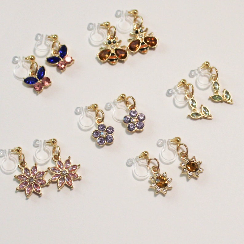 May include: A collection of clip-on earrings with various designs. The earrings feature gold-colored metal and colorful rhinestones in shapes like butterflies, bees, flowers, leaves, and suns. The earrings are displayed on a white surface.
