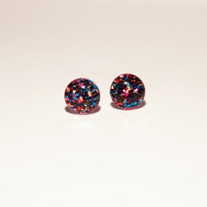 May include: A pair of round stud earrings with a glittery finish. The earrings are a mix of blue, pink, and gold glitter.