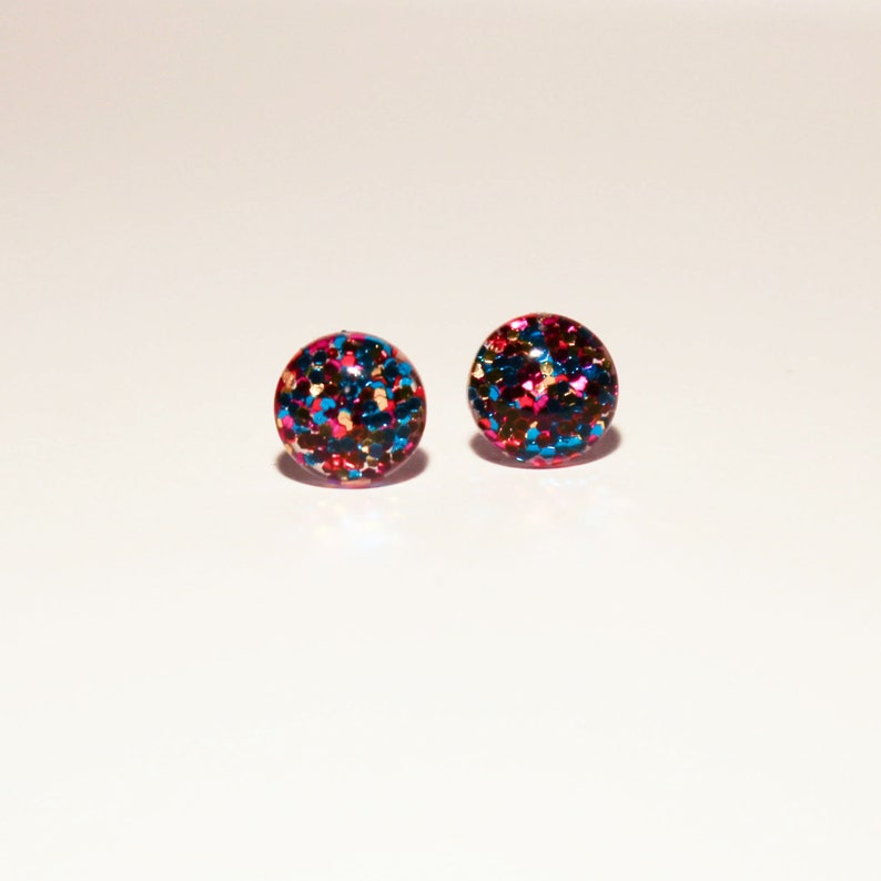 May include: A pair of round stud earrings with a glittery finish. The earrings are a mix of blue, pink, and gold glitter.