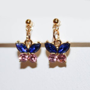 May include: A pair of gold-toned butterfly earrings. Each earring features a butterfly design with blue and pink gemstones. The earrings have a post and dangle design, suitable for pierced ears. The gemstones are faceted, adding sparkle.