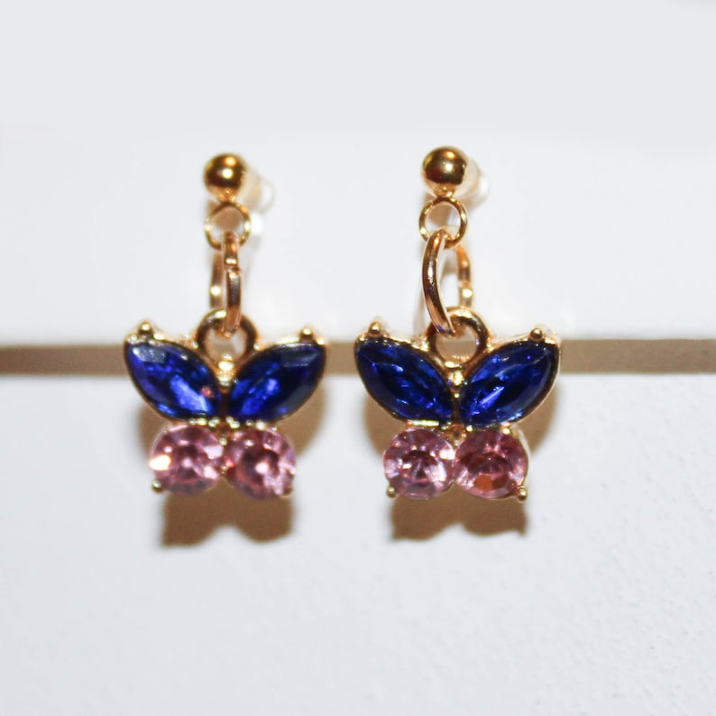 May include: A pair of gold-toned butterfly earrings. Each earring features a butterfly design with blue and pink gemstones. The earrings have a post and dangle design, suitable for pierced ears. The gemstones are faceted, adding sparkle.