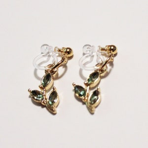 May include: A pair of gold-toned clip-on earrings featuring a leaf design. Each earring has three green, teardrop-shaped gemstones. The clip-on mechanism is clear plastic. The earrings are displayed against a white background.