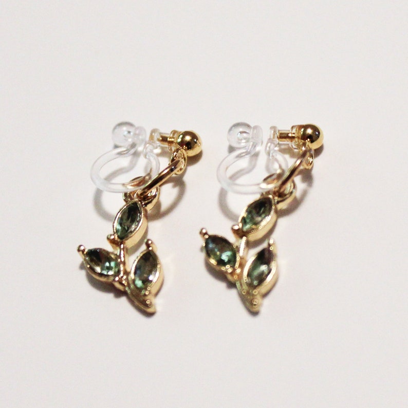 May include: A pair of gold-toned clip-on earrings featuring a leaf design. Each earring has three green, teardrop-shaped gemstones. The clip-on mechanism is clear plastic. The earrings are displayed against a white background.