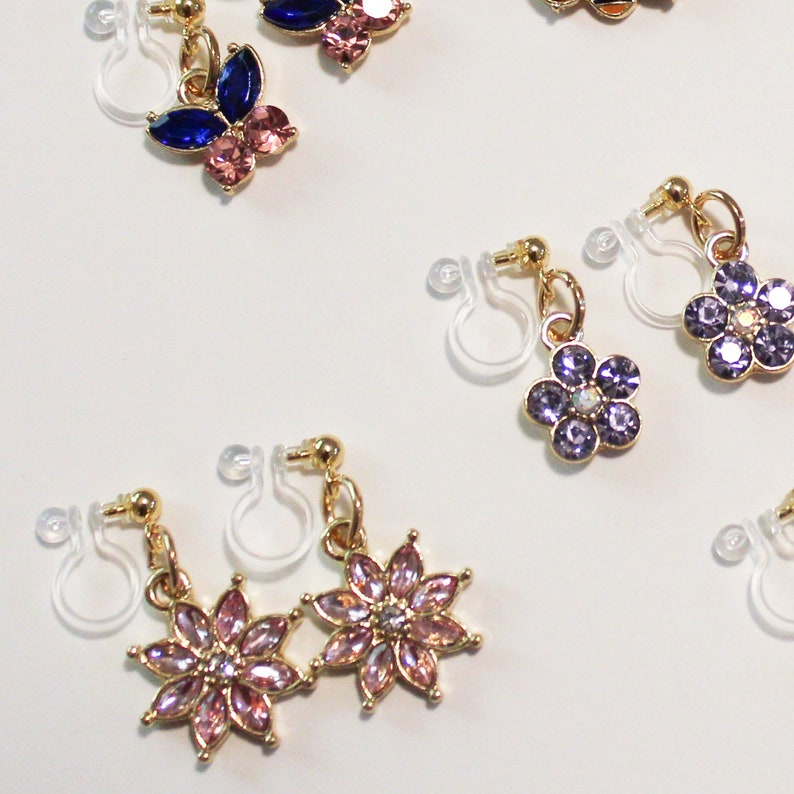 May include: A collection of clip-on earrings with gold-colored metal accents. The earrings feature various designs, including butterflies and flowers, adorned with pink, blue, and purple rhinestones. The earrings are displayed against a white background.
