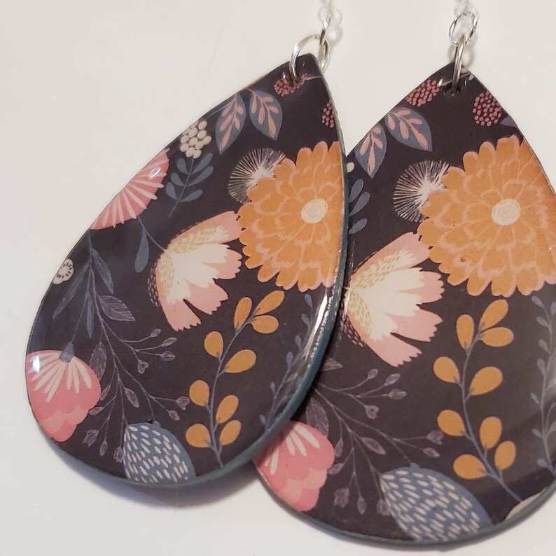 May include: A pair of teardrop-shaped earrings with a floral pattern. The earrings are black with pink, orange, and blue flowers. The earrings are made of resin and have silver hooks.