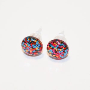 May include: A pair of small, round stud earrings with a colorful glitter design. The earrings are red, blue, yellow, and pink.