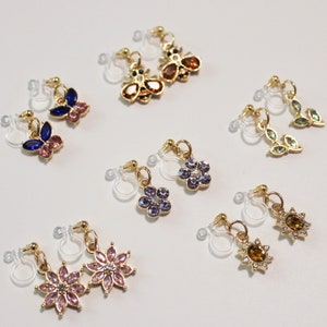 May include: A collection of clip-on earrings with various designs. The earrings feature gold-colored metal and are adorned with colorful rhinestones in shapes like butterflies, bees, flowers, leaves, and suns. The earrings are displayed on a white surface.