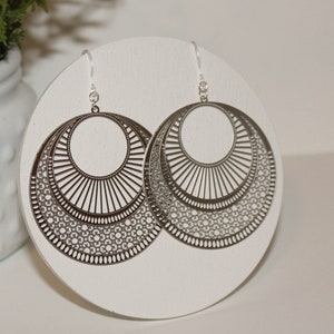 May include: A pair of silver-colored, layered hoop earrings with intricate cut-out designs. The earrings feature a circular shape with multiple concentric rings, each adorned with detailed patterns. The earrings are displayed on a white circular surface.