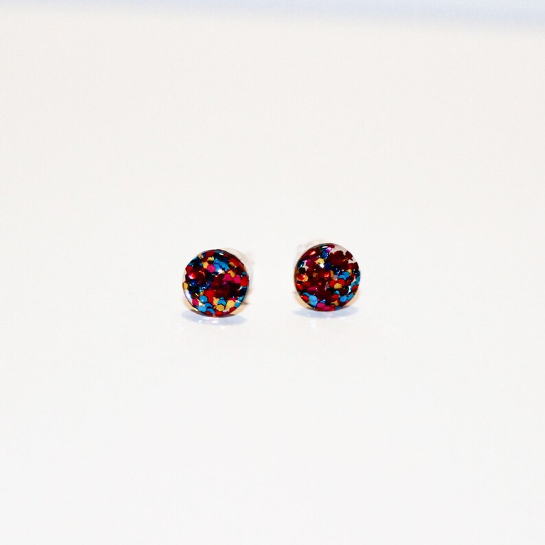 May include: A pair of small, round stud earrings with a red, blue, and gold glitter design.