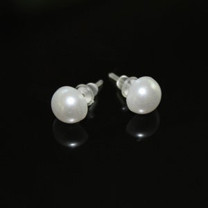 May include: A pair of white pearl stud earrings. Each earring features a round, lustrous pearl attached to a clear post with a plastic backing. The earrings are set against a black background, highlighting their simple elegance.