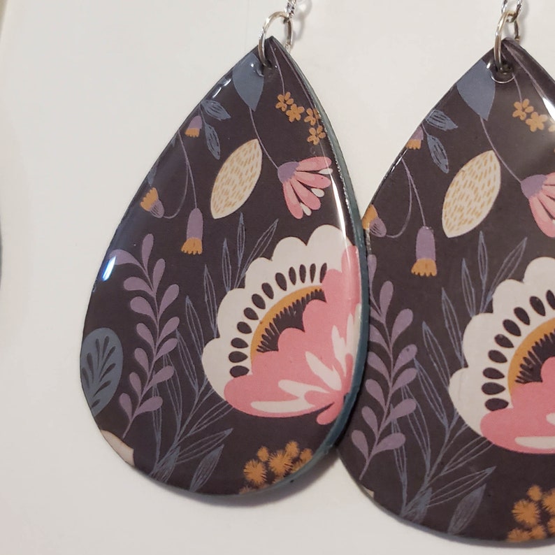 May include: A pair of teardrop-shaped earrings with a floral pattern. The earrings are black with pink, white, and yellow flowers. The earrings are made of resin and have a glossy finish.
