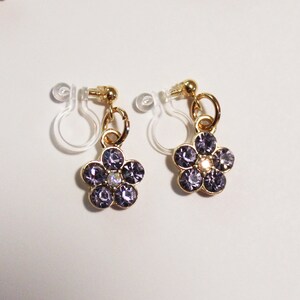 May include: A pair of gold-toned clip-on earrings, each featuring a flower design. The flowers are composed of purple rhinestones with a central iridescent stone. The earrings have a clear, plastic clip mechanism.
