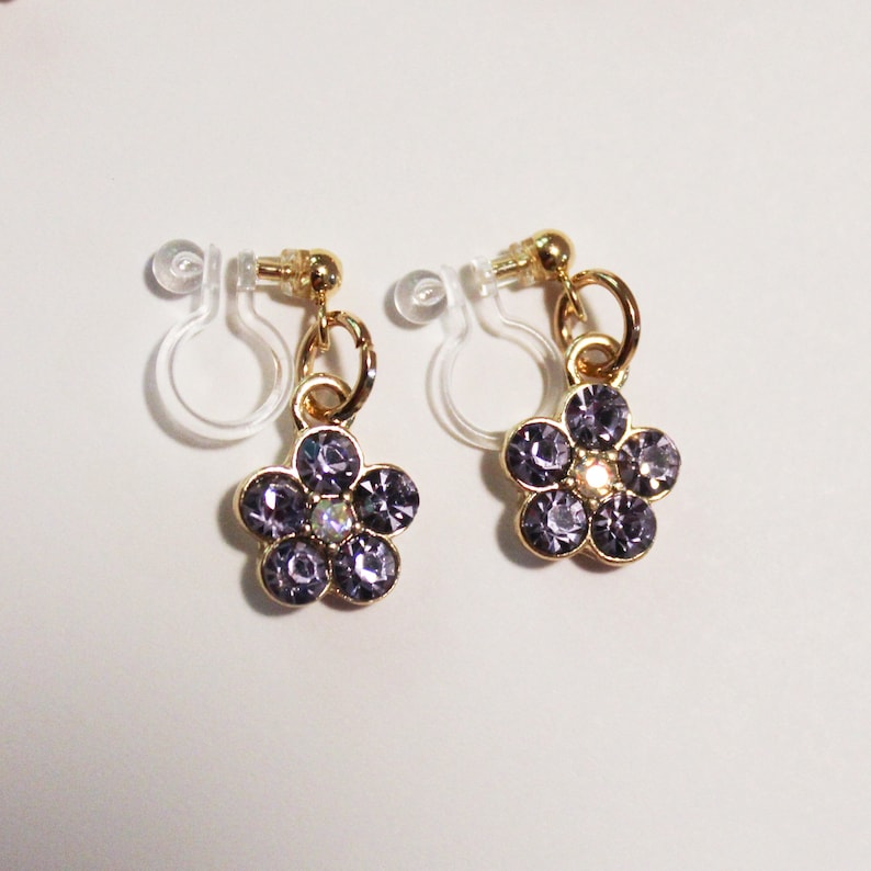 May include: A pair of gold-toned clip-on earrings, each featuring a flower design. The flowers are composed of purple rhinestones with a central iridescent stone. The earrings have a clear, plastic clip mechanism.