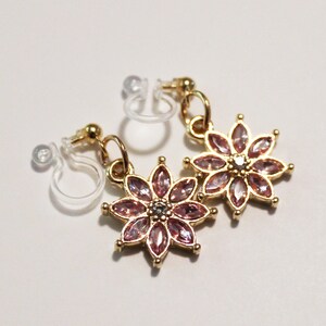 May include: A pair of gold-toned flower earrings with pink gemstone petals and a small, clear clip-on backing. The earrings are designed in a floral pattern, with each petal showcasing a faceted gemstone. The earrings are displayed on a white surface.