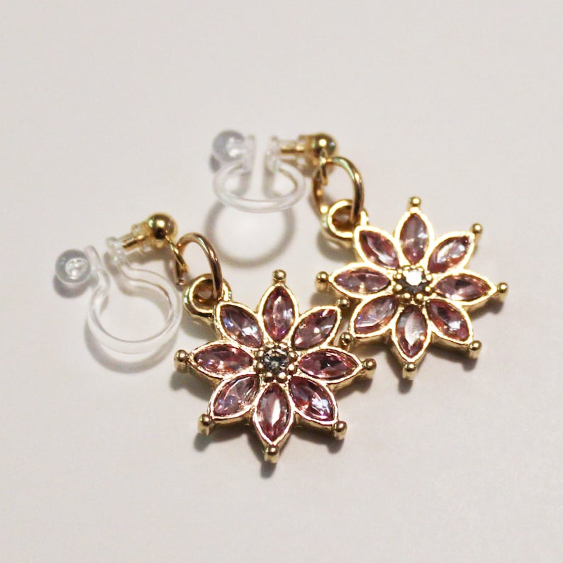 May include: A pair of gold-toned flower earrings with pink gemstone petals and a small, clear clip-on backing. The earrings are designed in a floral pattern, with each petal showcasing a faceted gemstone. The earrings are displayed on a white surface.