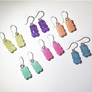 May include: A collection of colorful gummy bear earrings. Each pair features a translucent gummy bear charm in various colors, including green, purple, pink, orange, and blue, suspended from clear ear wires with gold-colored accents.