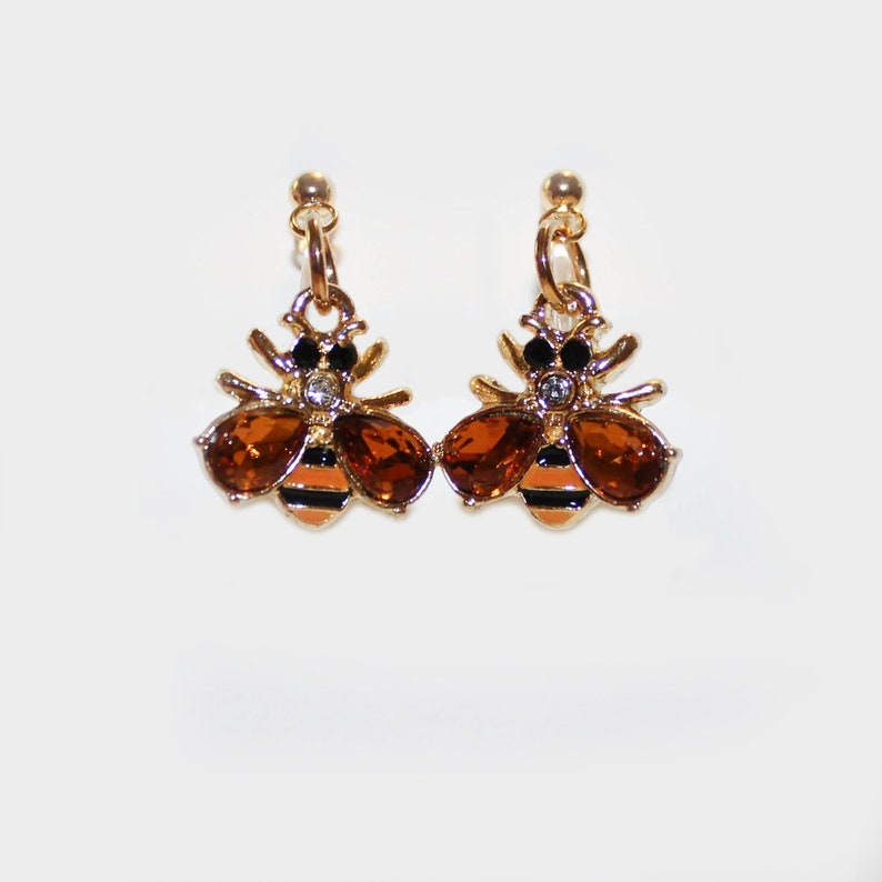 May include: A pair of gold-toned bee earrings. Each earring features a bee design with amber-colored gemstone wings, black and orange striped abdomen, and a small clear gemstone accent. The earrings have a post-back closure.