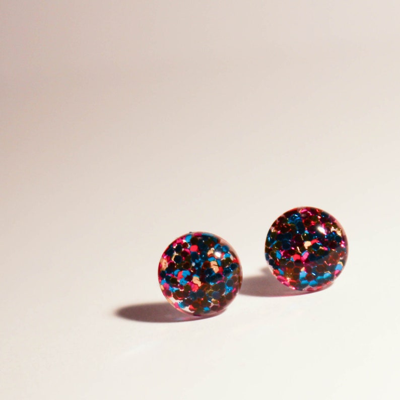 May include: A pair of round stud earrings with a glittery finish. The earrings are filled with blue, red, and gold glitter.