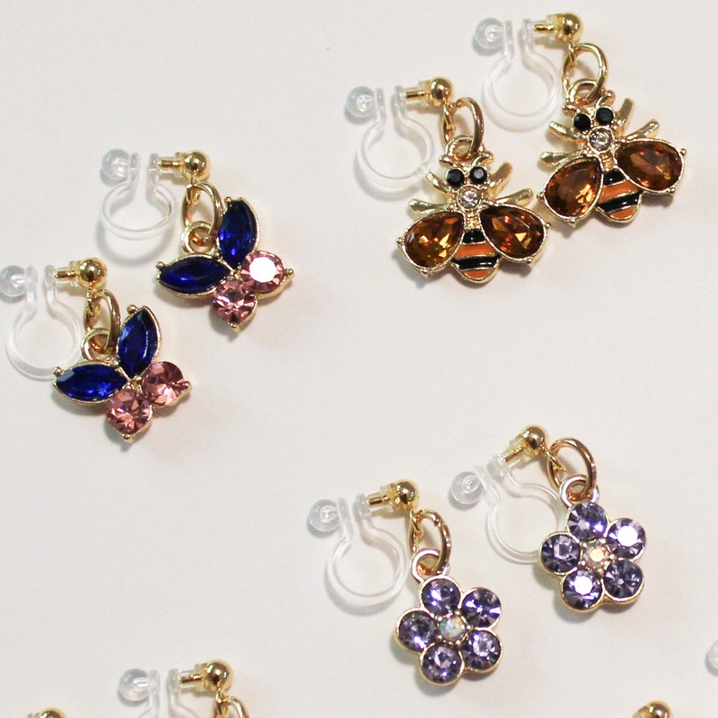 May include: A collection of clip-on earrings with gold-colored metal accents. The earrings feature various designs, including butterflies with blue and pink gems, bees with brown gems and black stripes, and purple flower designs. The earrings are displayed on a white surface.