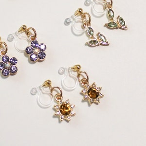 May include: A collection of clip-on earrings with gold-colored metal accents. The earrings feature various designs, including purple flower shapes, green leaf shapes, and sunburst shapes with amber-colored centers and clear crystal accents.