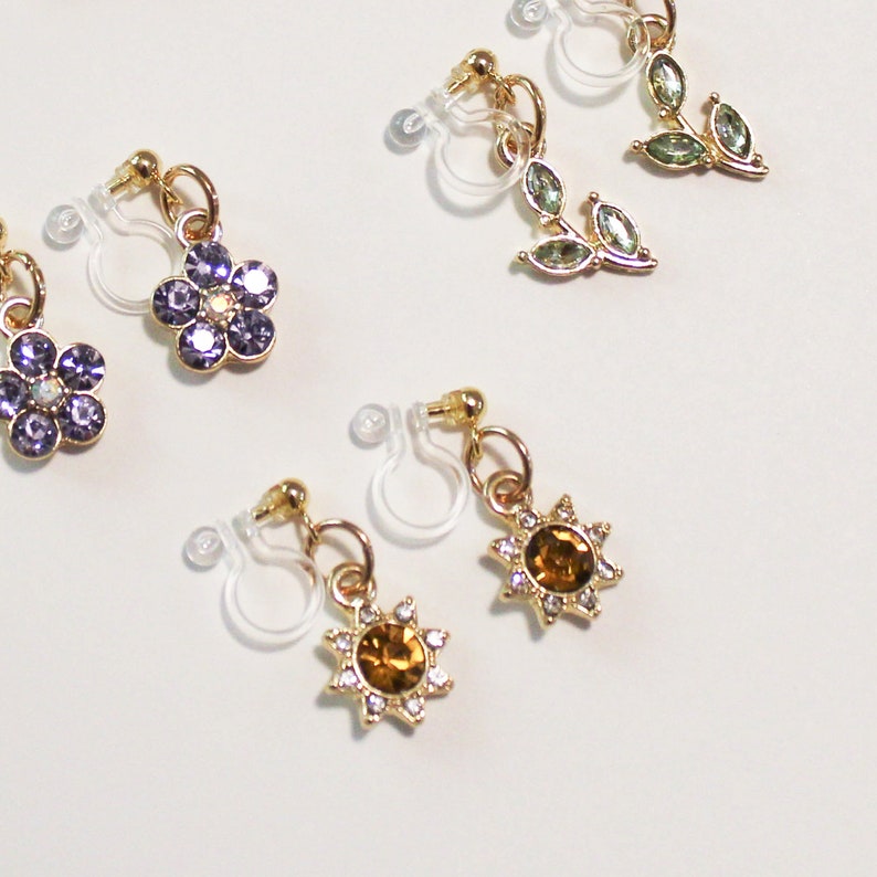 May include: A collection of clip-on earrings with gold-colored metal accents. The earrings feature various designs, including purple flower shapes, green leaf shapes, and sunburst shapes with amber-colored centers and clear crystal accents.