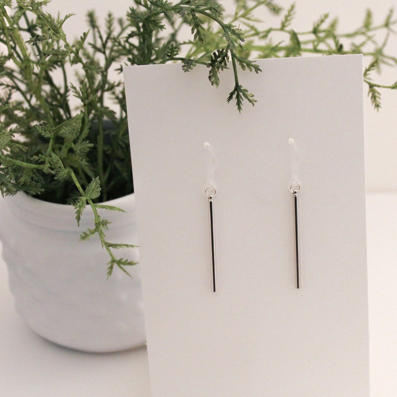 May include: A pair of silver dangle earrings with a thin, straight bar pendant. The earrings are displayed on a white card.