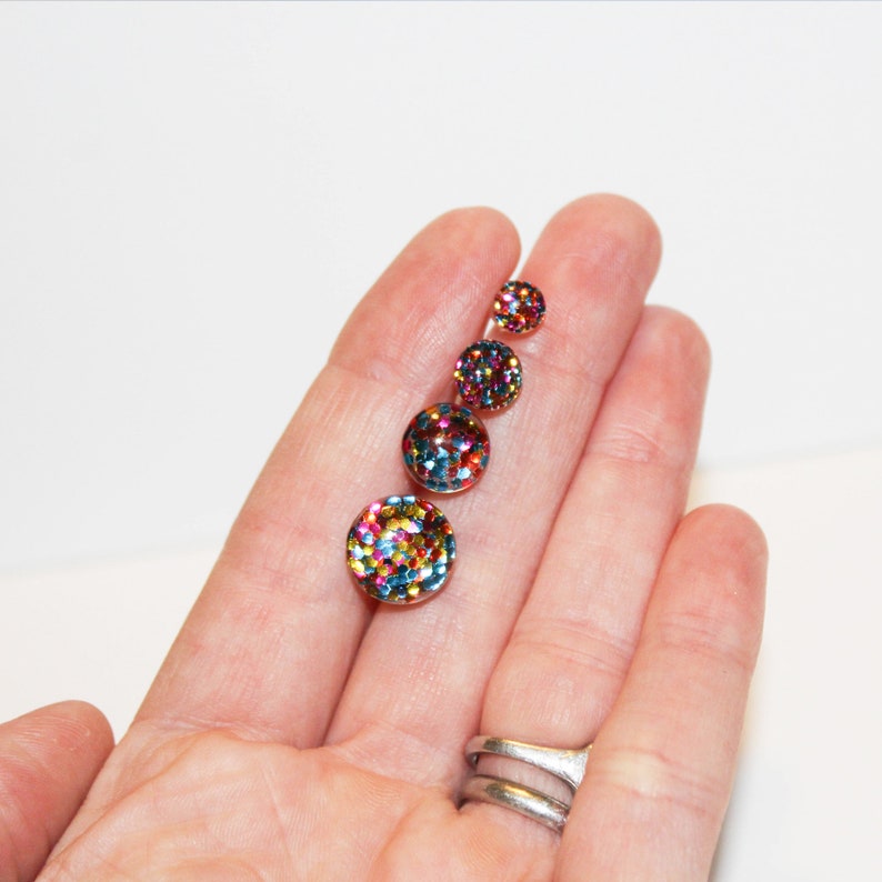 May include: Five round, colorful glitter-covered buttons in different sizes. The buttons are arranged in a line from largest to smallest on a hand.